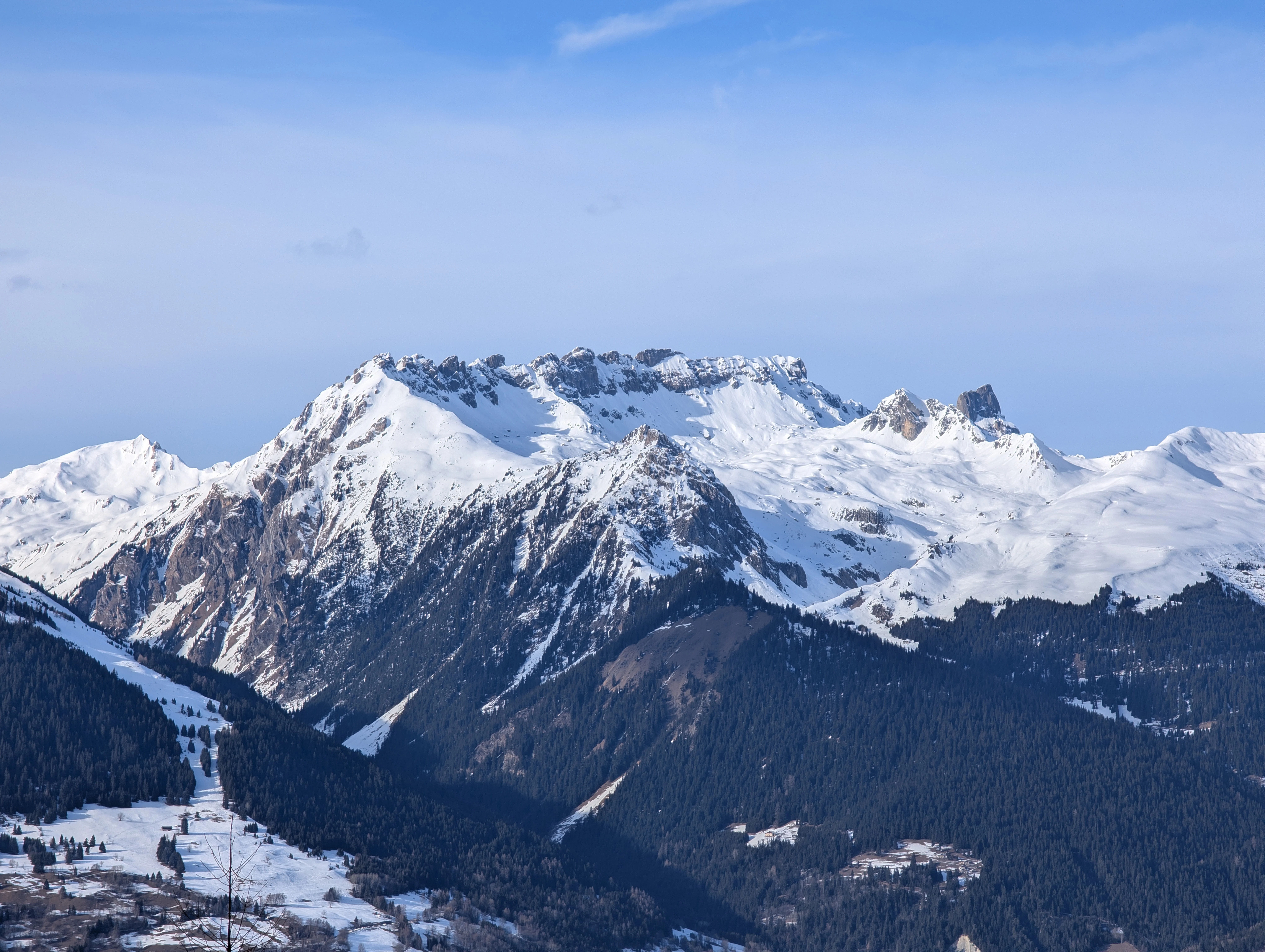 Snow-covered mountain peaks rise above forested slopes near Aime-la-Plagne, France, beneath a somewhat clear blue sky.